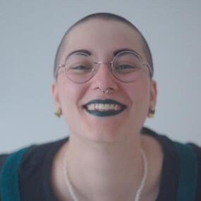 A picture of me, Lea, with a fresh buzzcut, black painted eyebrows, green lipstick, round glasses and a big smile.